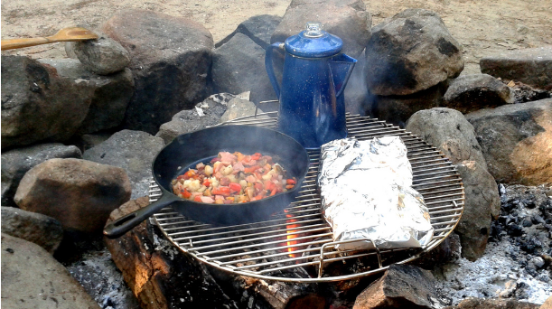 Food being cooked over a campfire, foil packed food and a cast iron skillet, and a kettle.